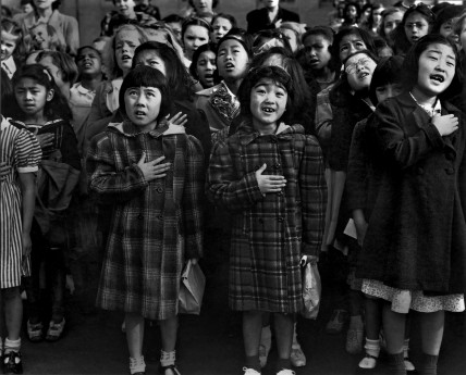Photo by Dorothea Lange_National Archives: Second-generation Japanese Americans, Helene Nakamoto Mihara, 7, left, and Mary Ann Yahiro, 7, center, recite the Pledge of Allegiance at the Raphael Weill School in San Francisco, CA, before being sent to the Topaz Internment Camp in Utah in April, 1942. Photo by Dorothea Lange/National Archives.